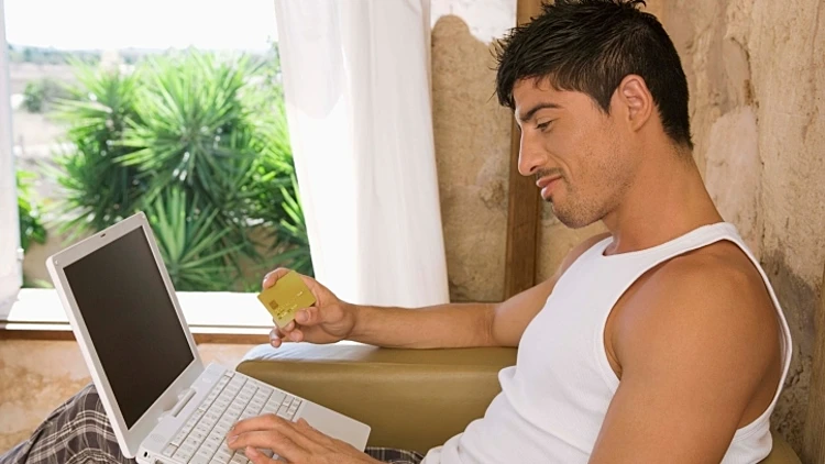 בחור יושב בגופיה על כורסה עם מחשב נייד מחזיק כרטיס אשראי