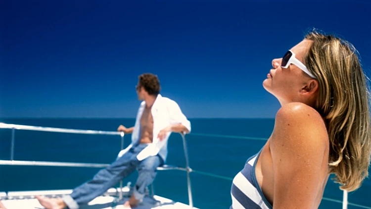 אישה בבגד ים ומשקפי שמש משתזפת על סיפונה של אוניה. הרחק רואים גבר עם חולצה פתוחה