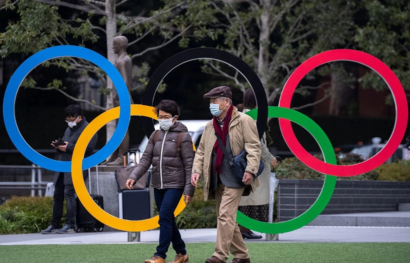 יפנים עוטים מסכות מפני קורונה מול סמל האולימפיאדה בטוקיו
